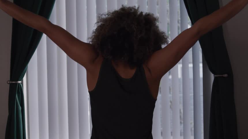 A woman with voluminous curly hair is shown from behind facing a window with vertical blinds she raises her arms above her head as if stretching or celebrating