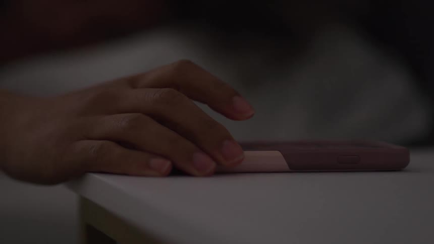 A young woman picks up her ringing phone placed beside the bed and answers the call while still lying on the bed