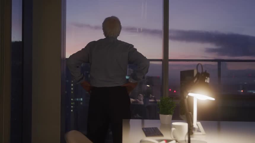 An older man standing with his hands on his hips looking at the city view from his office window