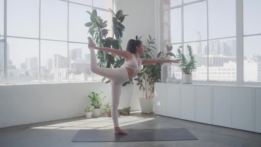 A woman practicing yoga in her apartment in front of windows with a view of the city