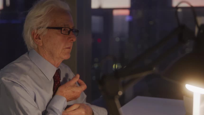An older man with white hair and glasses wearing a light blue shirt and a red tie adjusts the sleeve of his shirt while looking intently at something off screen with a desk lamp and blurred city lights in the background
