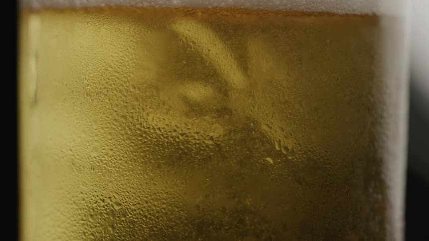 Condensation covers the outside of a glass of goldencolored beer as tiny bubbles slowly rise to the foam at the top