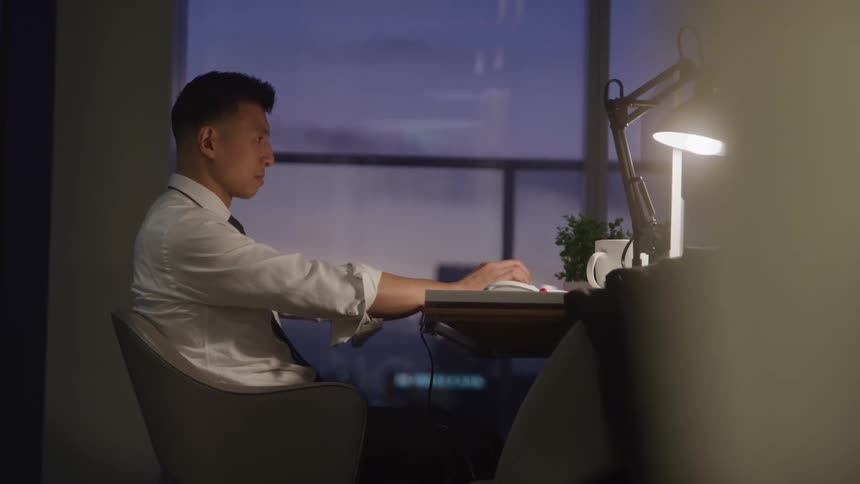 A young man sitting behind a desk in an office at sundown working on his laptop