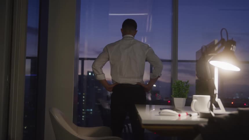 A businessman gazes out over the city from a high rise at dusk