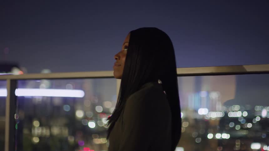 Woman smiles while standing outdoors near a clear barrier overlooking the city at night
