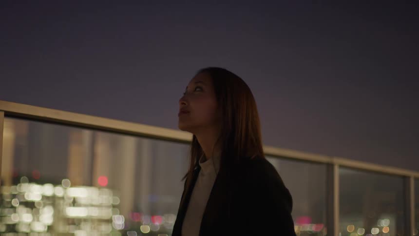 The video shows a woman standing outdoors at night looking upwards with a contemplative expression blurry city lights are visible in the background suggesting she is on a rooftop or balcony overlooking a city