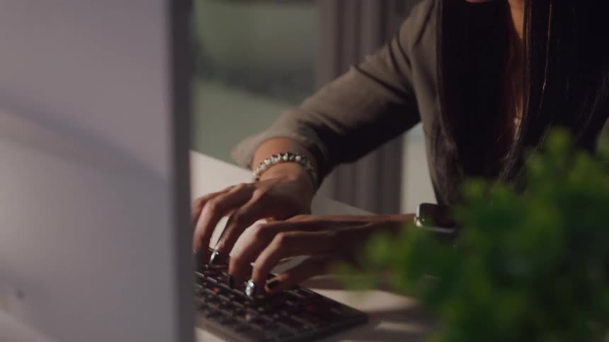 A woman with long dark hair is shown at a desk looking intently at a screen offcamera she is wearing a dark top or jacket and has a necklace visible