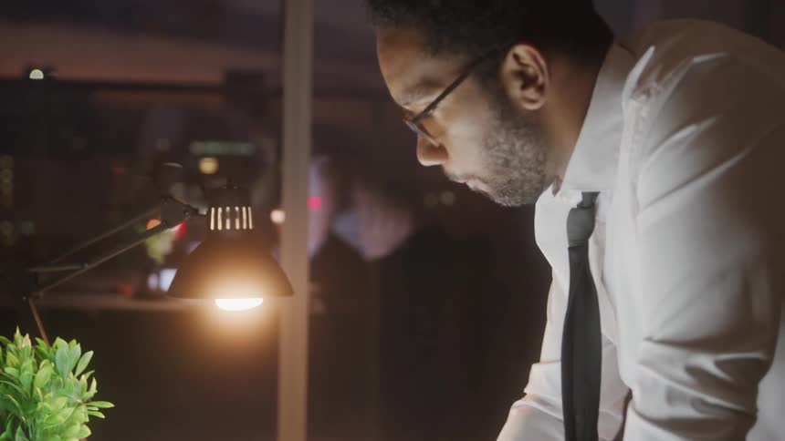 A focused black man wearing glasses a white shirt and a dark tie illuminated by a desk lamp