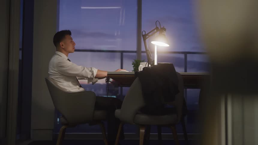 A man in a white shirt and tie at a desk illuminated by a bright lamp with a city view at dusk visible through the large windows behind him