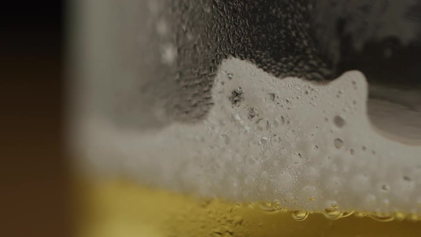 A static extreme closeup shot captures the lacy white foam and cold condensation clinging to the inside of a glass of goldencolored beer