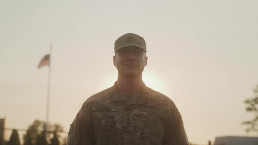 A military service member in uniform stands at attention silhouetted against a bright sky with an american flag visible in the background