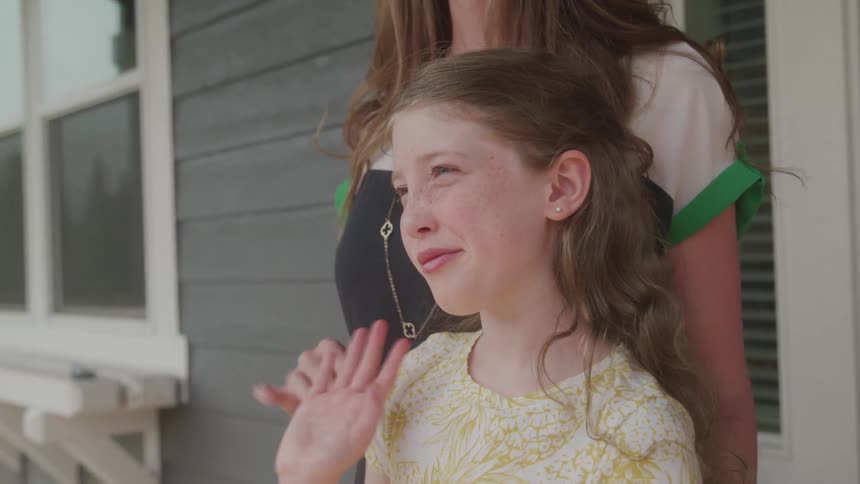The video features a closeup shot of a young girl with long reddishbrown hair standing outside a house with a woman the girl looks to the side and waves her hand seemingly at someone offcamera