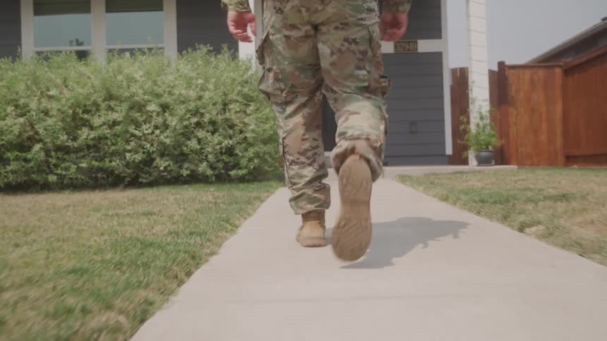 The video shows a person in military camouflage trousers and boots walking along a concrete pathway towards a house the camera focuses on their legs and feet as they walk with a suburban home and landscaping visible in the background