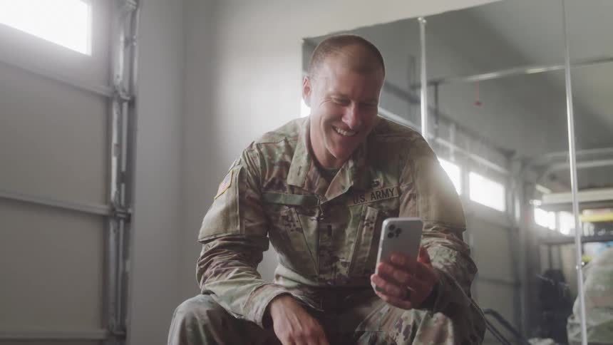 The video shows a man in a us army uniform sitting down and having a video call on his phone he appears to be smiling and waving at the camera suggesting a cheerful conversation the setting looks like an indoor space with large windows or garage doors