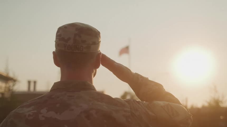 A person in military fatigues and a camouflage cap with the name young visible on the back stands facing away from the camera they are saluting with a bright sunset or sunrise and a flagpole in the background