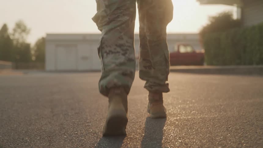 A pair of feet possibly wearing boots is shown walking on a paved surface during what appears to be late afternoon or early morning judging by the warm low sun the scene is somewhat industrial or urban with blurred buildings in the background