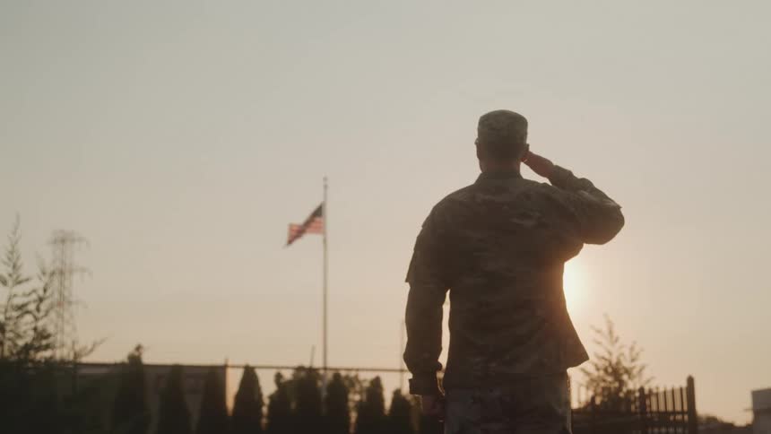 An american soldier salutes while standing in front of an american flag outdoors at sunset