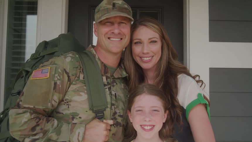 The video shows a family of three consisting of a man in military uniform a woman and a young girl smiling happily as they stand together in front of a house the man is wearing a camouflage uniform with a large backpack and a us flag patch is visibl