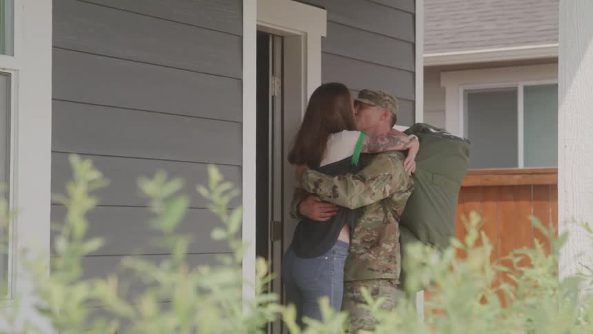 A woman opens the door to her house and kisses a man wearing a uniform