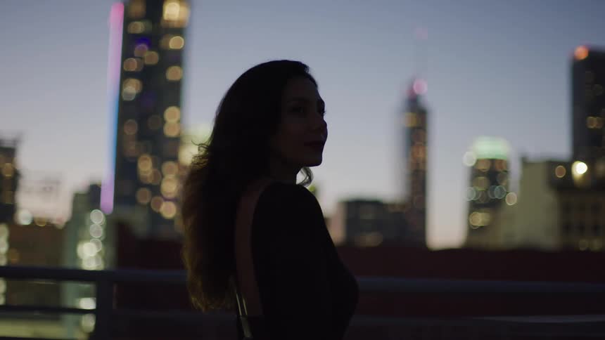 A young woman on top of a building roof at sundown with the cityscape in the background featuring a series of skyscrapers and highrise buildings