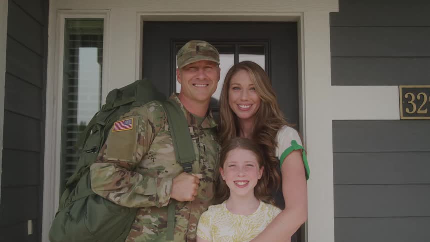 A soldier in camouflage uniform and a cap carrying a large backpack stands with a woman and a young girl in front of a house all three are smiling and looking at the camera suggesting a family reunion or departure