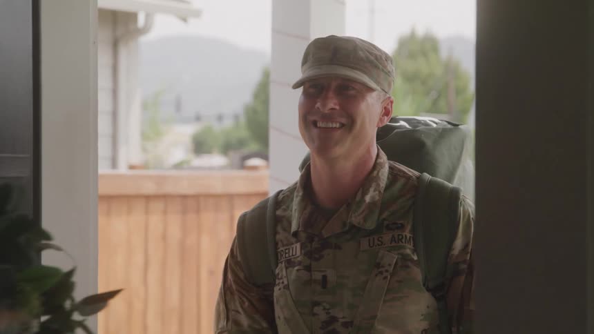 A woman with long hair opens a dark door revealing a man in a us army uniform with a backpack who smiles warmly at her they embrace suggesting a reunion