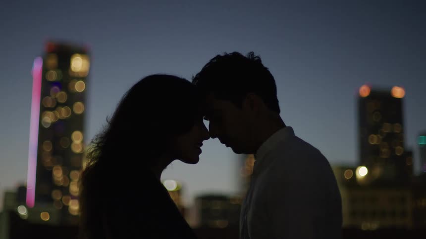The video shows two individuals in silhouette with their foreheads gently touching framed against a nighttime cityscape with blurred lights they stand close appearing to share an intimate moment outdoors