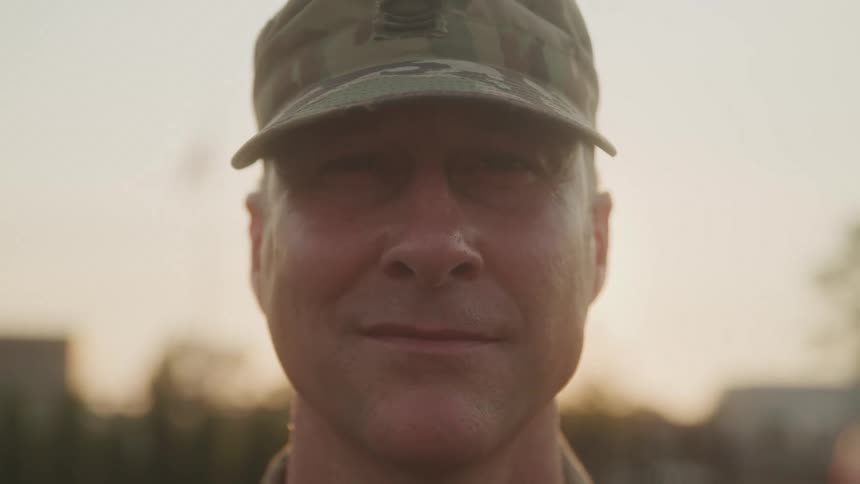 A soldier in uniform stands confidently outdoors with the sun setting behind him