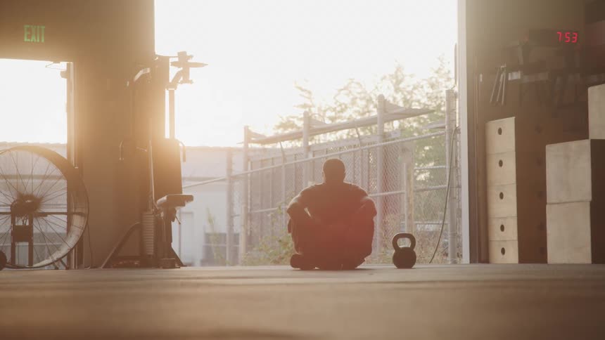 The video shows a person sitting on the floor in a gym or warehouse facing away from the camera towards a bright open entrance a kettlebell rests on the floor to their right the lighting is strong from the background creating a silhouetted effect