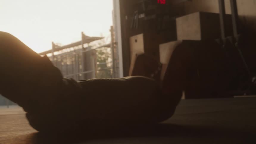 A man seen in silhouette performs sit ups on a floor with bright light from an open doorway in the background illuminating dust particles in the air and gym equipment stacked nearby