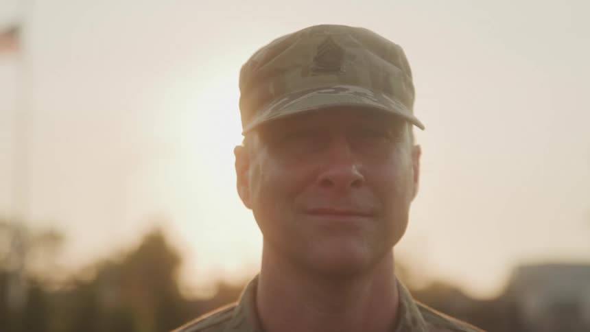 An adult male in military uniform wearing a camouflage cap looks directly at the camera with a subtle smile as bright sunlight shines from behind him