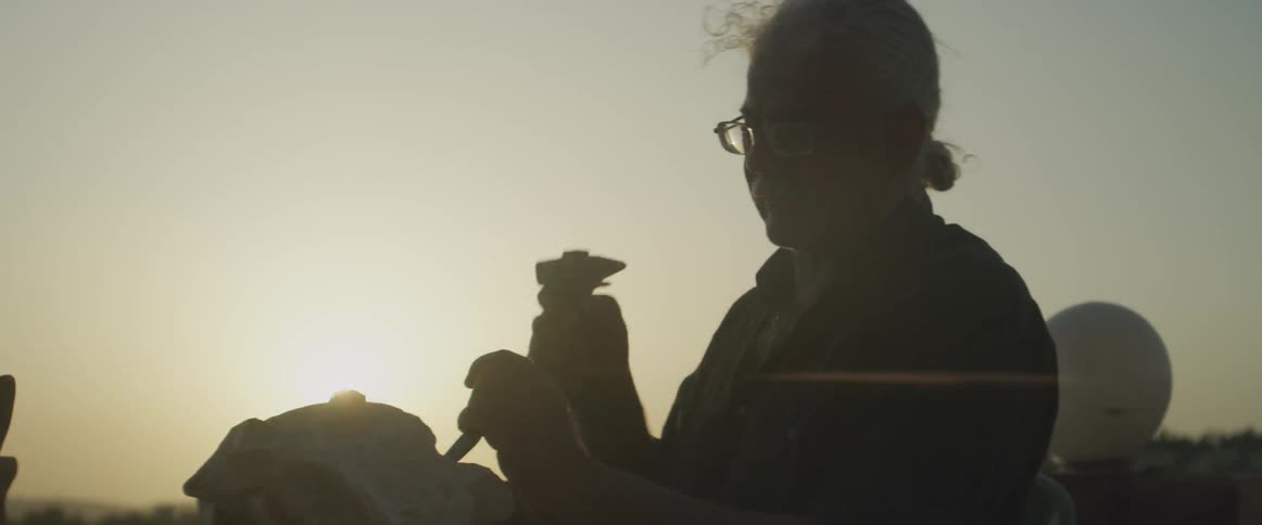 An elderly man works with a hammer and chisel outdoors