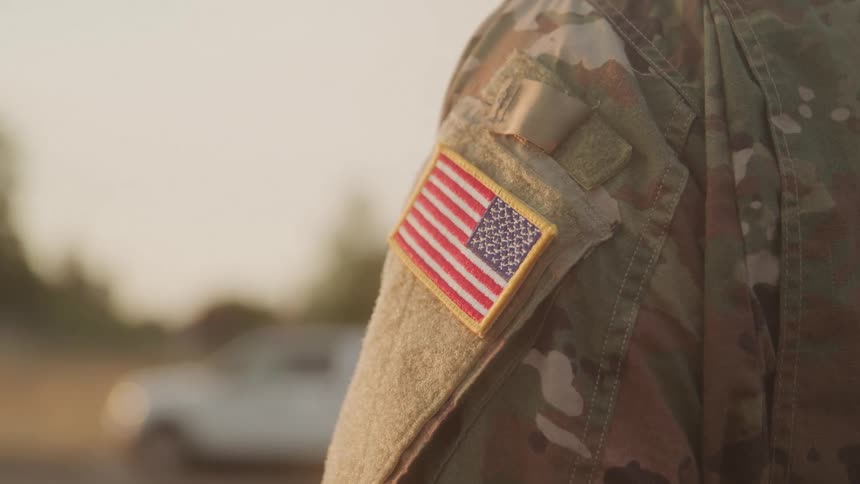 The video shows a closeup of a military uniform sleeve with an american flag patch prominently displayed the background is blurred suggesting an outdoor setting in daylight