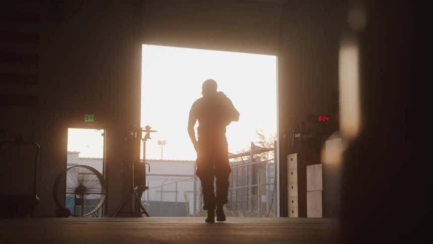The video shows a silhouetted figure walking towards a bright open doorway carrying a duffel bag or similar item over their shoulder the strong backlighting from the doorway possibly indicating sunrise or sunset creates a dramatic effect within what