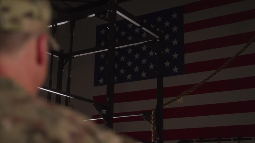 The video shows a person in military uniform from behind saluting a large american flag the flag is displayed prominently on a wall and theres what appears to be a gym or fitness rig in front of it