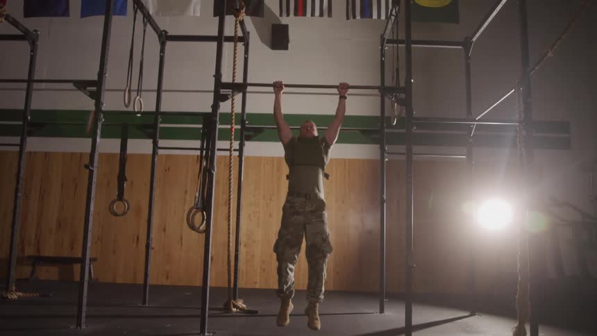 A man in military fatigues performs pull ups on a metal bar in a gym with various workout equipment and flags visible in the background