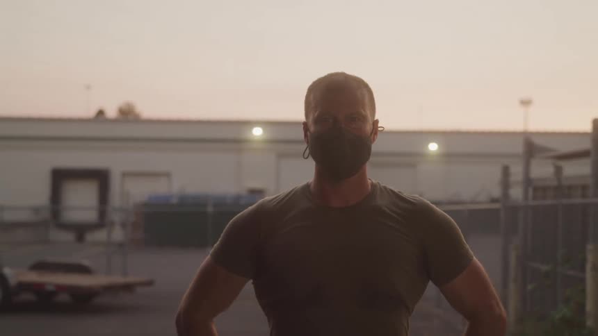 A man wearing a nose mask standing outdoors at dusk with both hands on his waist