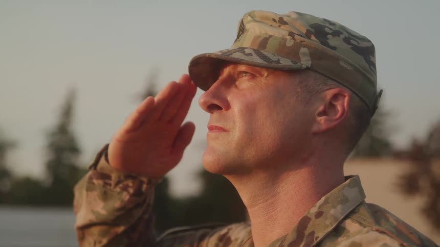 The video shows a closeup of a man in military camouflage wearing a cap as he brings his hand to his forehead in a salute he is looking off into the distance with a serious expression and the background is slightly blurred with hints of green