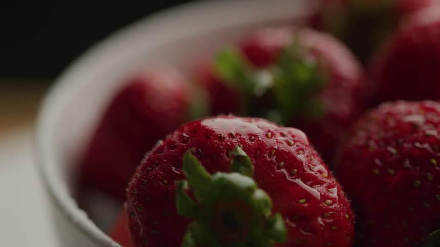 A bowl of fresh wet red strawberries with green caps is shown in a close up highlighting their juicy texture