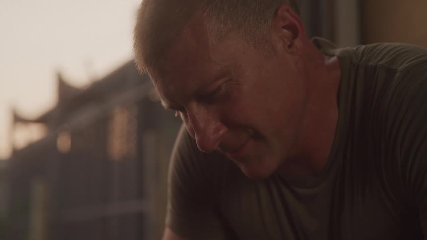 A mans face showing him looking down with a focused or contemplative expression illuminated by warm soft light from the upper lef