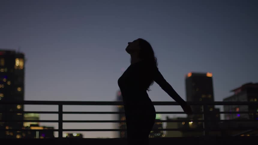 The video shows the silhouette of a woman standing on a rooftop or balcony at dusk with city buildings visible in the background she raises her arms and looks upwards seemingly enjoying the open space and evening sky