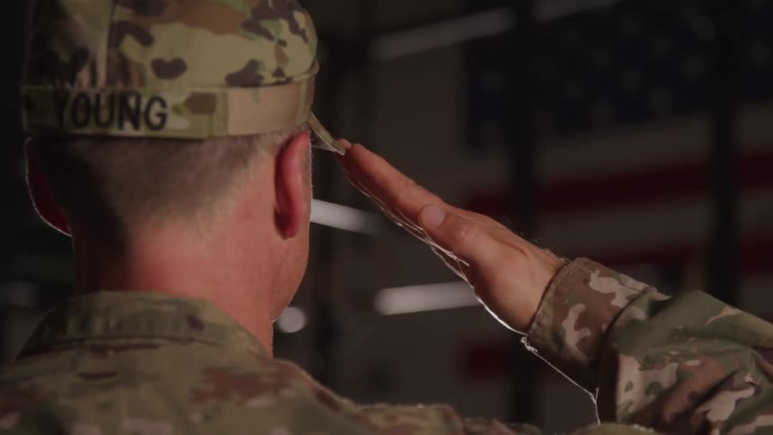 Soldier in camouflage uniform salutes with the right hand with a blurred united states flag in the background