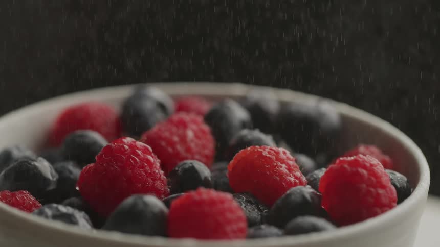 Fresh raspberries and blueberries sit in a white bowl as water gently sprinkles down