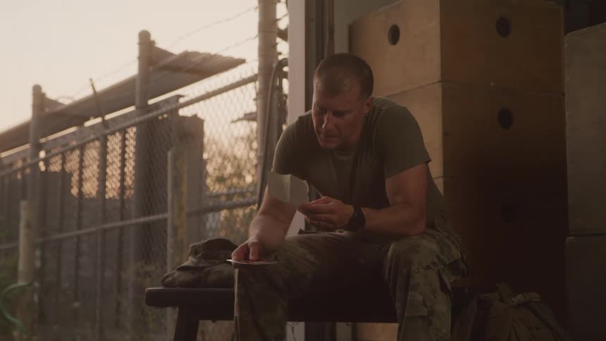 The video shows a man in what appears to be military fatigues sitting outdoors and looking intently at a small object in his hands possibly a photo or letter he has a serious and then slightly smiling expression as he interacts with the item in the ba