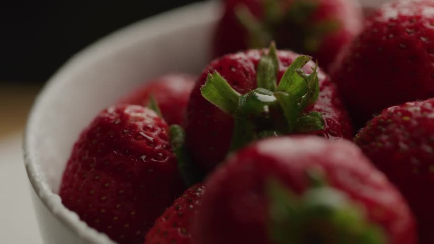 Fresh piled ripe strawberries fill a white bowl glistening with droplets of moisture