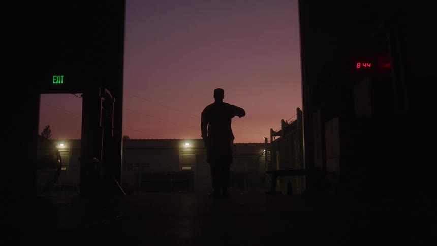 A male figure in silhouette stands in a large doorway looking out at a dusky sky raising his hand to his forehead in a salute with an illuminated exit sign and a digital clock displaying 844 visible on the dark walls