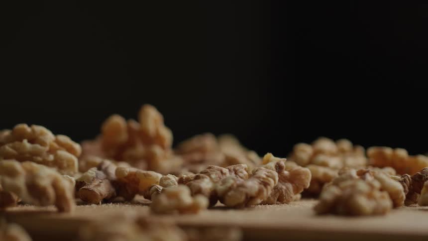 A pile of shelled walnuts rests on a light colored wooden surface against a dark out of focus backdrop
