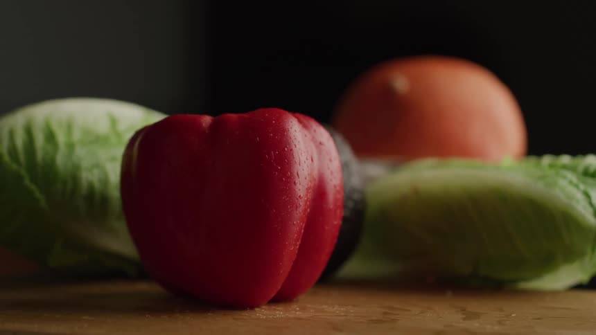 A fresh red bell pepper is dropped in slow motion onto a wet wooden surface coming to rest in the center of a colorful arrangement of lettuce an onion and other vegetables against a dark background