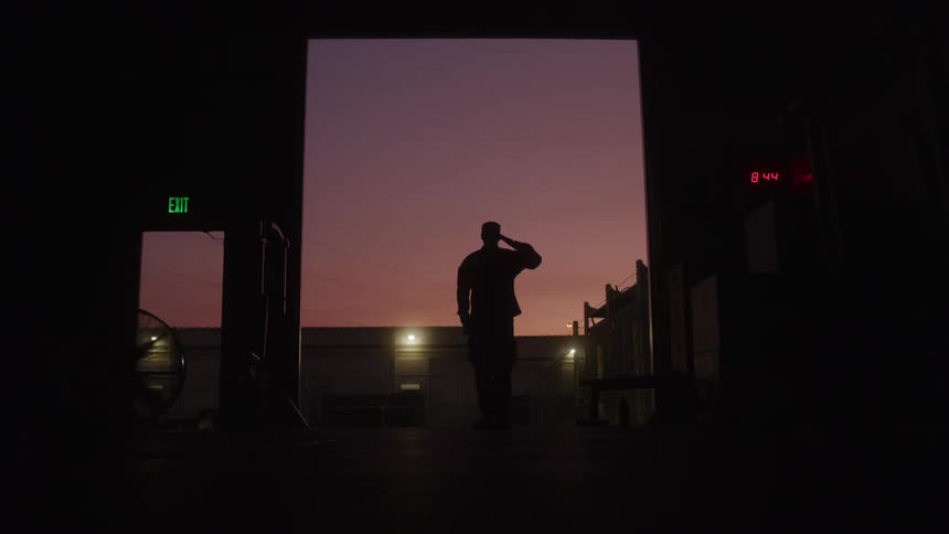 Man silhouetted by the red tinted sky stands in an open loading doorway on a loading dock and salutes