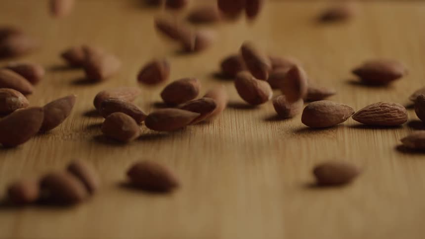 In a slowmotion closeup a continuous shower of raw almonds tumbles and bounces onto a lightcolored wooden cutting board gradually forming a scattered pile